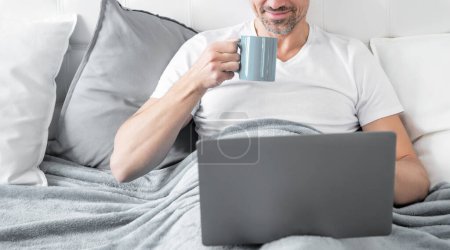 positive mature man working on laptop in bed with coffee.