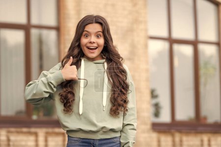 Surprised teenage girl in casual hoodie giving thumb gesture blurry outdoors, copy space.