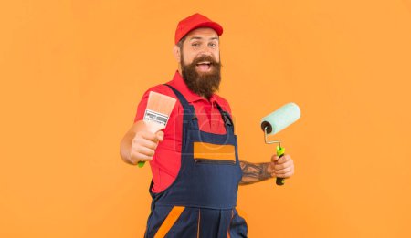 fröhliche bärtige Mann Hausmaler in Arbeitskleidung halten Farbroller und Pinsel und auf gelbem Hintergrund.