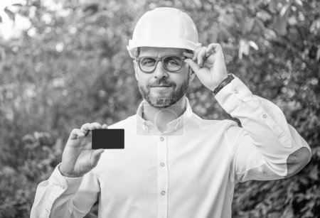 Hombre supervisor en hardhat mostrando la tarjeta de contacto en blanco al aire libre, espacio de copia.