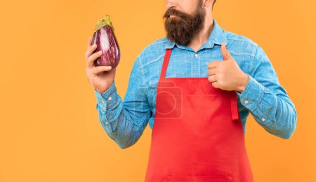 Serious man crop view in red apron giving thumb gesture to eggplant yellow background, greengrocer.