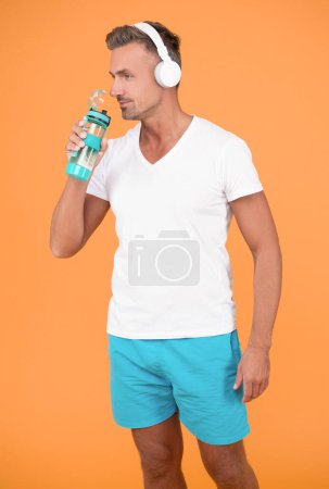 fröhlicher sportlicher Mann mit Kopfhörern trinkt Wasser. sportliche Mann trinken Wasser isoliert auf gelbem Hintergrund. Sportlicher Mann trinkt Wasser im Studio Sportler trinken nach dem Training Wasser.