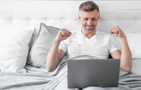 happy mature man working on laptop in bed.