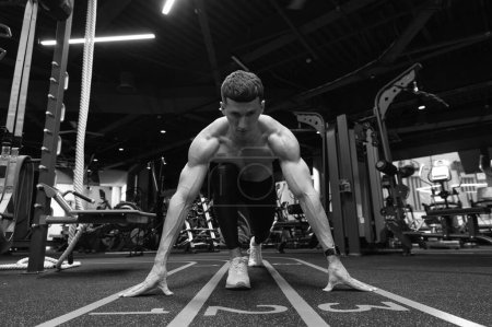 Athletic man in crouching position in fitness gym. Athlete with front knee over and hands on starting line in gym. Running training in gym. Run workout. Fitness and sport.