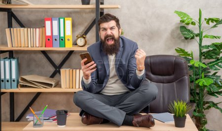 Excited man with smartphone exult in victory making winning gesture sitting cross-legged on office desk, succeed.