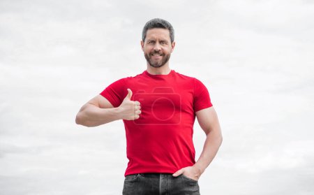 happy man in red shirt outdoor on sky background. thumb up.