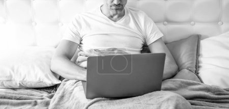 Reifer Mann mit Brille arbeitet am Laptop im Bett.