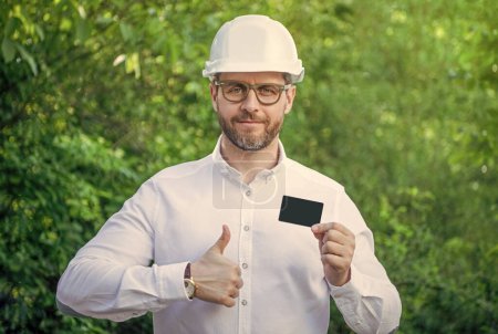 Man foreman in hardhat giving thumbs up showing blank contact card outdoors, copy space.