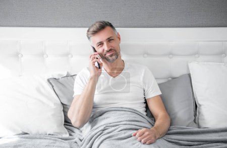 mature man smile in bed speaking on phone.