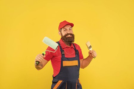 heureux homme barbu peintre en vêtements de travail tenir rouleau de peinture et pinceau sur fond jaune.
