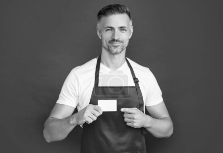 smiling mature man in apron with copy space on card. point finger.