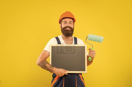 smiling man decorator in helmet and work clothes hold paint roller and blackboard with copy space on yellow background.