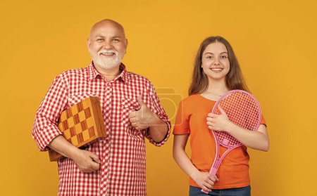 glad teen girl with grandfather with badminton racket and chess on yellow background.