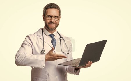 smiling doctor offering emedicine in studio. doctor presenting emedicine on background. photo of emedicine and doctor man with laptop. doctor promoting emedicine isolated on white.