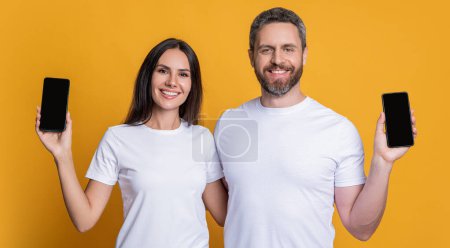 Man and woman showing phone app. Family blog. Product proposal. Advertisement copy space. Couple presenting phone screen. Couple showing smartphone isolated on yellow. Touchscreen technology.