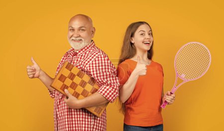 glückliches Teenager-Kind mit Großvater mit Badmintonschläger und Schach auf gelbem Hintergrund. Daumen hoch.