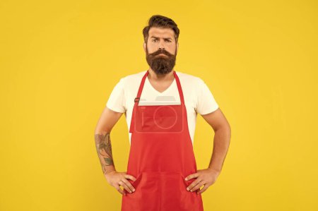 Confident serious man with arms akimbo in red apron yellow background, household.
