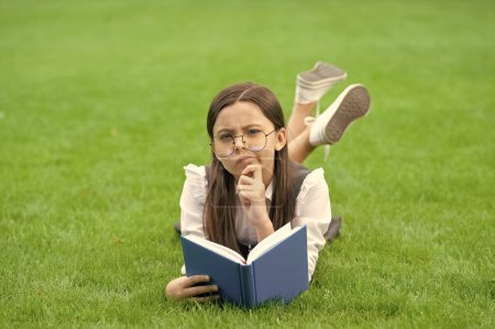 Téléchargez les photos : Pensant adolescent fille lecture livre couché sur l'herbe après l'école, l'éducation. École et éducation. - en image libre de droit