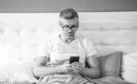 mature man in bed check email on phone wearing eyeglasses.