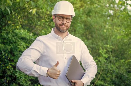 Architect man in hardhat giving thumbs up gesture holding laptop outdoors.