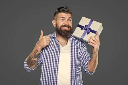Téléchargez les photos : Mature barbu gars avec boîte cadeau sur fond gris. pouce levé. - en image libre de droit