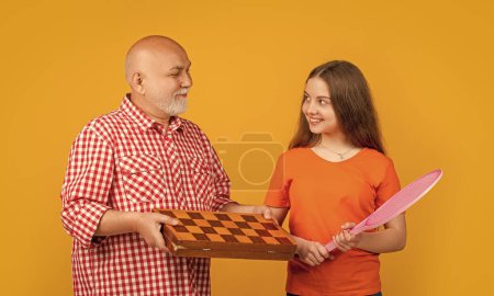 chica feliz con abuelo con raqueta de bádminton y ajedrez sobre fondo amarillo.