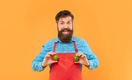 Happy man in apron holding fresh limes citrus fruits yellow background, greengrocer.