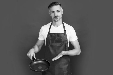 mature man chef in apron with frying pan on red background. presenting product.