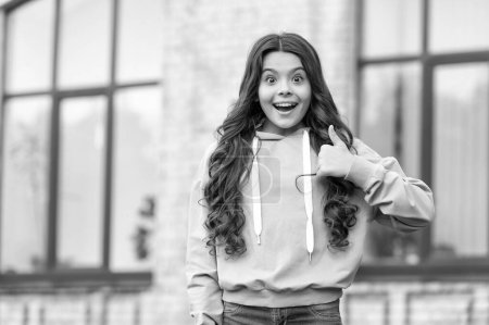Surprised teenage girl in casual hoodie giving thumb gesture blurry outdoors, copy space.