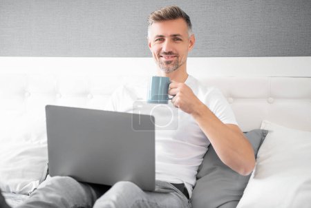 Téléchargez les photos : Happy mature man using computer in bed with coffee. - en image libre de droit