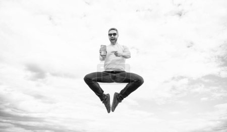 Erstaunter Mann springt und präsentiert Smartphone auf Himmelshintergrund.