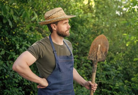 Serious rural man in farmers hat and gardening apron looking into distance holding garden shovel natural outdoors.