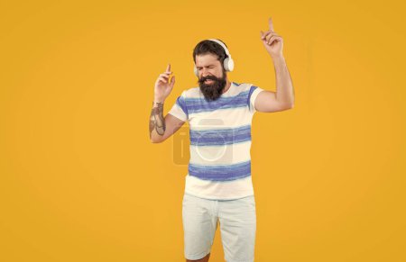Téléchargez les photos : Hipster man écouter de la musique dans des écouteurs isolés sur jaune. pointant du doigt. homme dans les écouteurs hipster écouter de la musique. Prise de vue en studio d'un hipster avec écouteurs. écouter de la musique. dispositif moderne de l'homme. - en image libre de droit