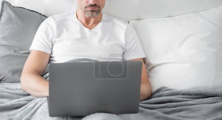 mature man working on laptop in bed. business online.