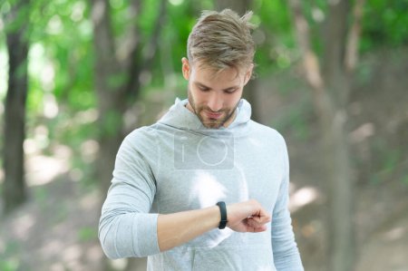 Man tracking fitness results dressed in sportswear outdoor. Man runner with fitness gadget. Smartwatch for fitness. Sporty fit man with sport equipment checks time on smartwatch. Check running time.