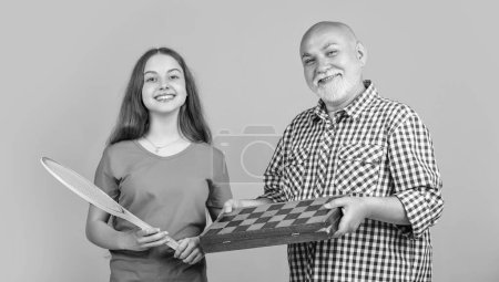 Photo for Smiling teen child with grandfather with badminton racket and chess on yellow background. fathers day - Royalty Free Image