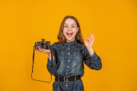 Happy teenager showing OK ring gesture holding photo camera yellow background, photographer.