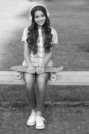 Téléchargez les photos : Joyeuse adolescente avec planche à roulettes dans un casque. adolescent fille avec planche à roulettes à l'extérieur. adolescent fille avec skateboard écouter de la musique. adolescent fille avec skateboard en plein air. - en image libre de droit