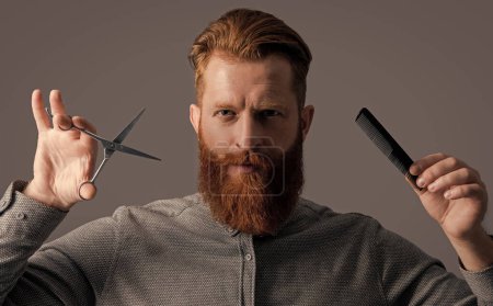 handsome barber man with scissors do hairstyle. man has beard and hairstyle in studio. man hold barber scissors and comb. barber with hairstyle. barber man making hairstyle isolated on grey background