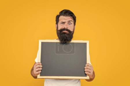 disappointed bearded man hold blackboard for advertising. studio man advertising. bearded man advertising on blackboard with copy space. bearded man advertising with blackboard isolated on yellow.