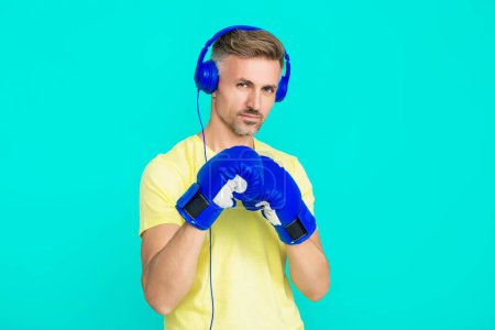 Mature man boxer in headphones. Sport man boxing. Strong man listen music. Sportsman in gym. Boxer in sportswear. Exercising and punching. Sportsman in boxing gloves isolated on blue. Achievement.