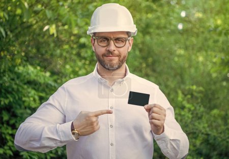 Businessman man in hardhat pointing finger at blank business card outdoors, copy space.