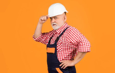 serious aged man builder in helmet on yellow background.