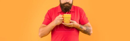 Bearded man crop view smelling hot beverage in cup yellow background, coffee.