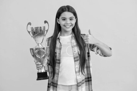 teen girl receive award in studio, thumb up. teen girl accept award on background. proud teen girl with champion cup award. teen girl hold her award champion cup isolated on yellow.