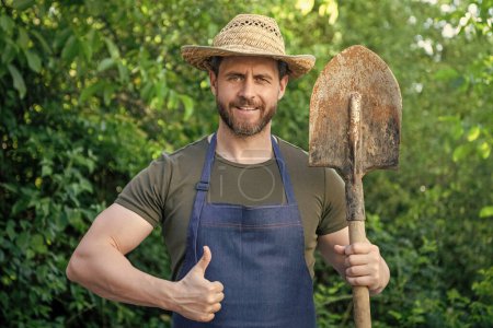 Happy gardener man in gardening apron and farmers hat with garden spade giving thumb gesture natural outdoors.