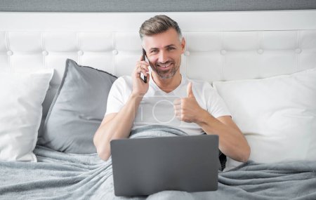 mature man speaking on phone and working on laptop in bed. thumb up.