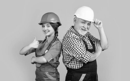 happy teen child and grandfather builder in helmet on yellow background. thumb up.
