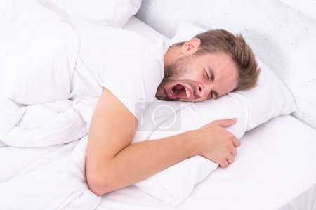 Yawning early in the morning. Asleep young man sleeping. Resting peacefully in comfortable bed. Lying with closed eyes. Man sleeping at night. Sleepy man lying on bed sleeping at white bedroom.