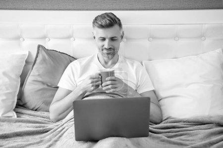froher reifer Mann, der im Bett mit Kaffee am Laptop arbeitet.